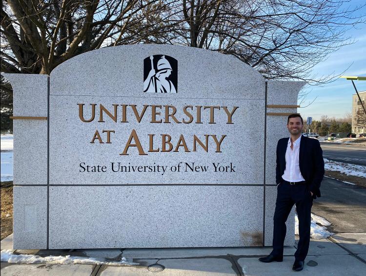 O aluno Patricio está, de pé, sorrindo ao lado de uma parede de concreto que contém o logo da SUNY Albany